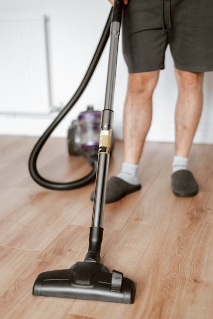 A professional cleaner from East Sheen Carpet Cleaners wearing a white protective suit, face mask, and blue gloves is performing deep cleaning on a light beige carpet in a bright, modern living room. 